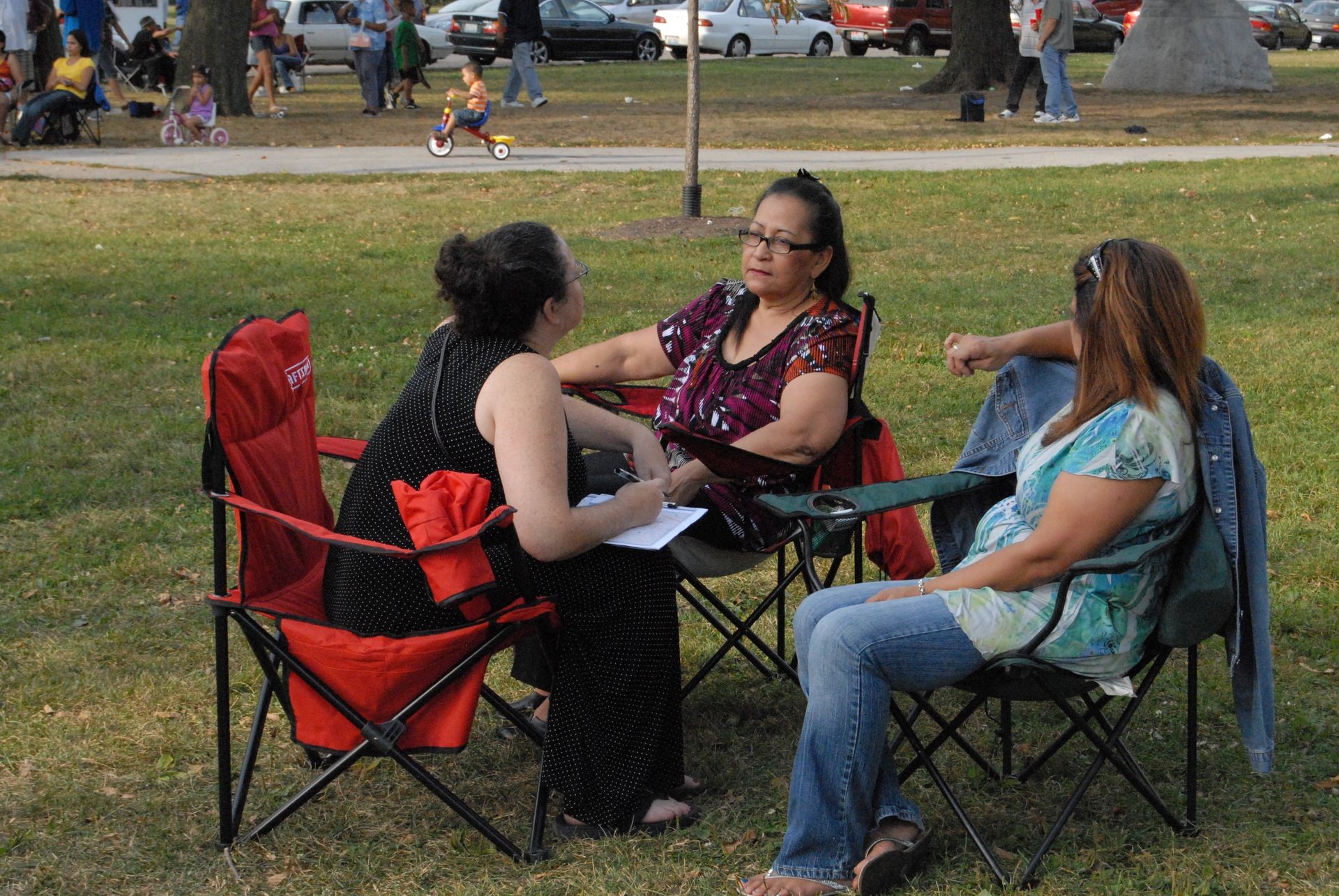 Three women sitting on folding chairs in a park, engaged in conversation.