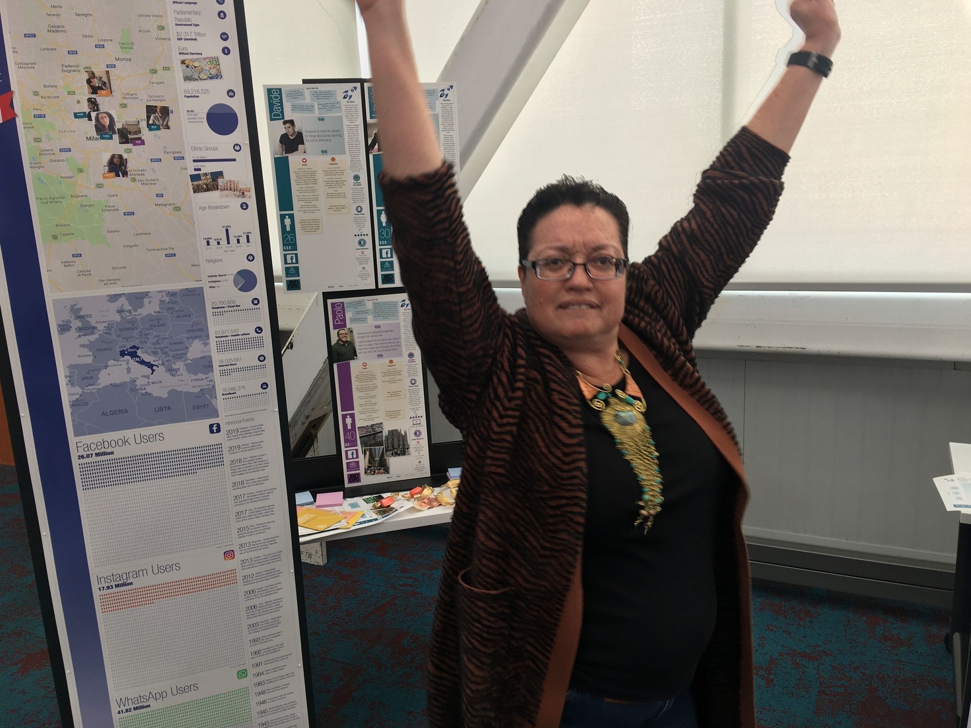 Person with raised arms standing next to informational posters and maps in an exhibition hall.